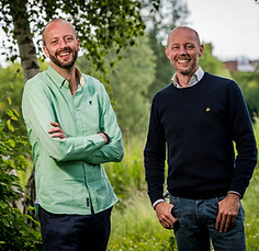 Two smiling men standing outdoors in front of green foliage and trees, one wearing a light green shirt with folded arms and t