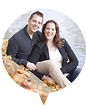 Smiling couple sitting on a leaf-covered path beside a body of water, dressed in fall clothing.