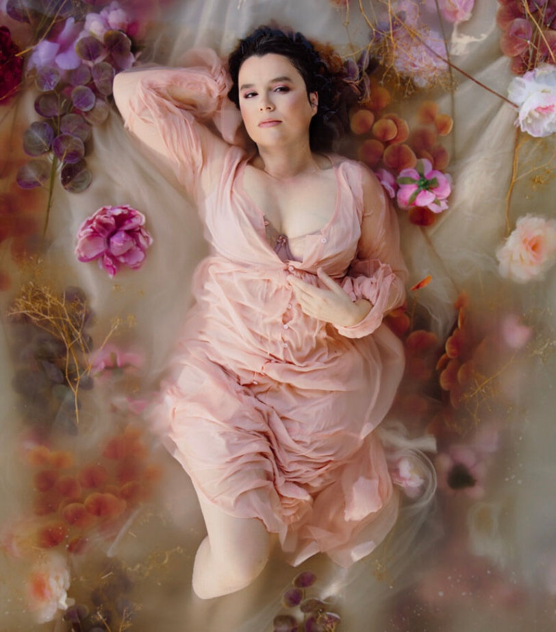 A woman in a pink dress lies in water surrounded by floating flowers. The serene setting has soft colors, creating a dreamlike mood. Bay Area Boudoir by Liza Head.