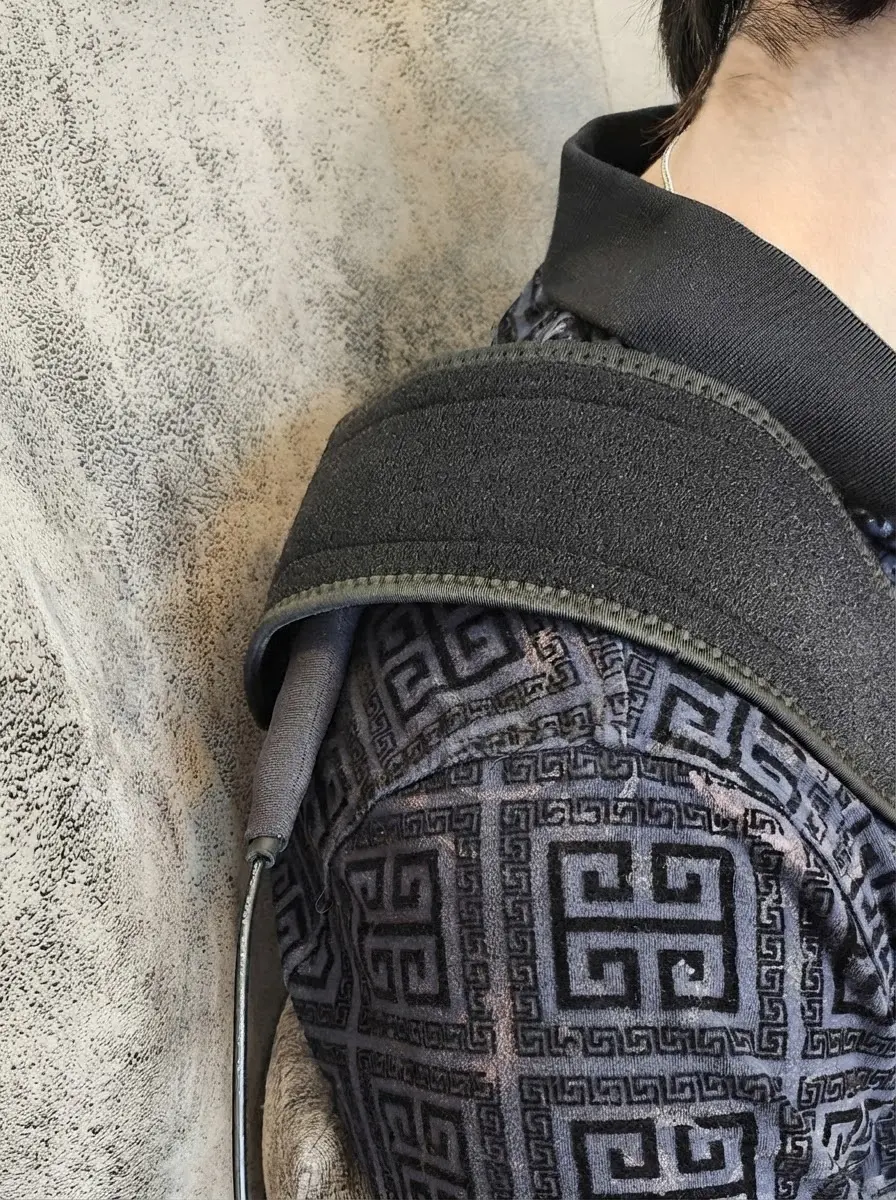 Studio photography detail of a person wearing ornate patterned clothing and a shoulder strap. The intricate geometric design suggests cultural or historical significance.