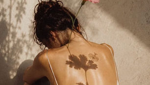 Mulher sentada em silêncio, de costas, sob a luz do sol, com uma flor rosa nos cabelos e sombras de flores sobre o corpo.