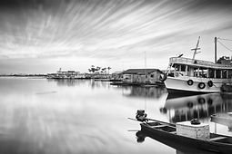 Fotografia em preto e branco de uma comunidade ribeirinha no Amazonas, capturada com longa exposição; rio suavizado como superfície lisa, céu em movimento, casas flutuantes e barco branco na margem; atmosfera minimalista e contemplativa.