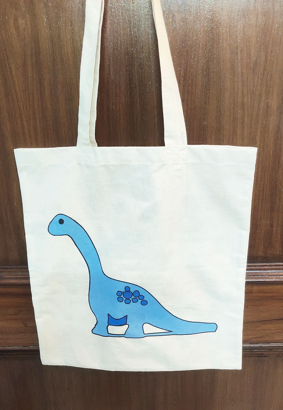 Thumbnail: White tote bag with a print of a blue dinosaur. the bag is hanging on a  door