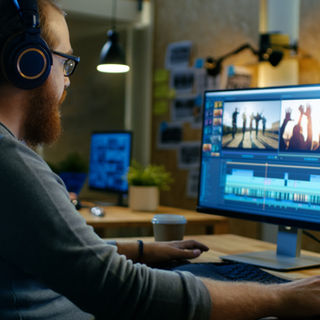 A video editor wearing headphones works on a project using editing software on dual monitors.