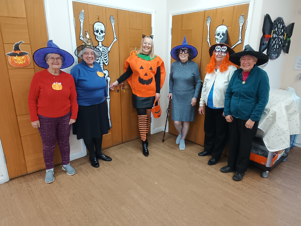 Six women in Halloween costumes, smiling in a festive room with skeletons and pumpkins on wooden doors. Hats and orange accents stand out.