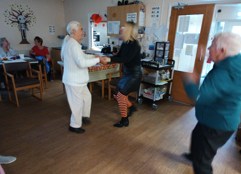 Two people dance in a room, one wearing striped leggings. A skeleton decoration and seated people are in the background. Mood is joyful.
