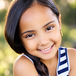 Outdoor headshot of a smiling young girl with dark hair wearing a striped top.