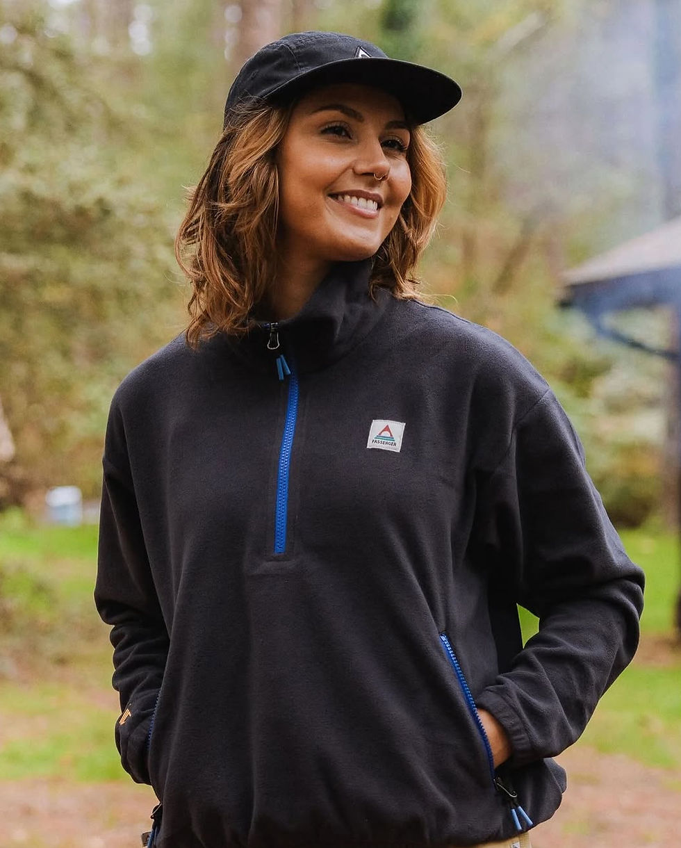 Female model in a cap wearing a black outdoor jacket smiling in the woodland.