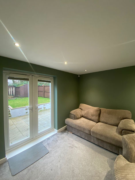 A modern living room in Brighouse, West Yorkshire, featuring deep green painted walls, a white ceiling with recessed lighting, and a wooden coffee table against a freshly decorated backdrop. The room is finished with grey carpeting and has a glass door leading to an outdoor patio area.