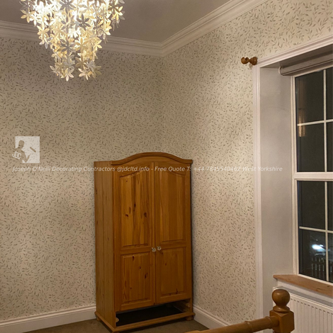 A professionally redecorated bedroom featuring soft floral wallpaper, clean white crown molding, a wooden bed frame, and a decorative hanging light fixture. The room exudes a cozy and elegant atmosphere, perfect for a comfortable living space. Project completed by JDC Ltd in West Yorkshire