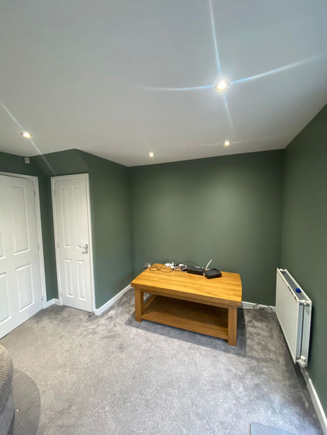 A stylish living room in Wakefield, West Yorkshire, featuring deep green painted walls, a white ceiling with recessed lighting, and a soft grey carpet. The room is furnished with a large beige corduroy sofa and a matching swivel chair. French doors open onto a well-maintained garden, allowing natural light to brighten the space. A modern, cozy, and professionally decorated interior.