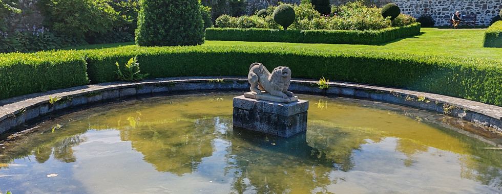 Stone fountain with a lion statue in a circular pond, surrounded by manicured hedges and a stone wall. Sunlit garden with lush greenery.