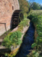 Old stone mill wall with a dark, vertical water wheel beside a narrow stream, surrounded by lush green plants and trees.
