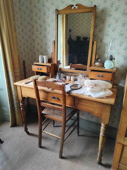 Antique wooden vanity with a mirror, brushes, and perfume bottles. Floral wallpaper and a window curtain create a vintage, cosy setting.
