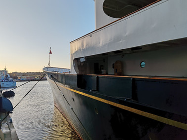 Britannia docked at sunset, dark blue hull with a gold stripe. Small boat P627 in the background. Calm water, clear sky, relaxed mood.