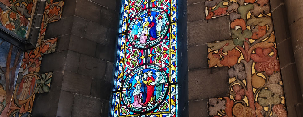 Stained glass window in a stone wall, depicting colourful biblical scenes. Intricate painted murals with vibrant patterns surround the window.