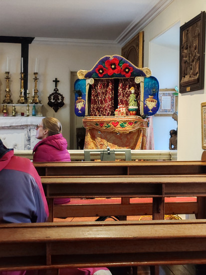A woman in a pink jacket sits on a pew in a chapel facing an ornate altar with colourful decorations and candles, creating a serene atmosphere. On display is a puppet show set.
