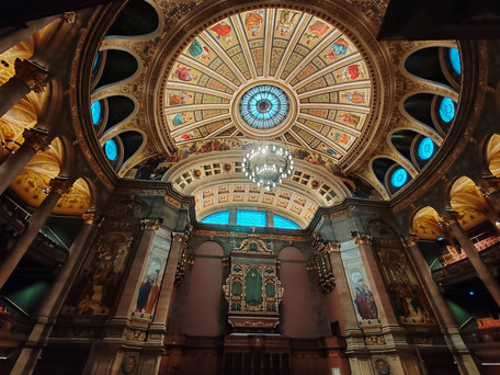 Ornate hall interior with a detailed dome ceiling, colourful stained glass, chandeliers, and religious artwork on walls. Elegant and grand.