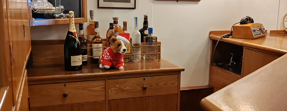 Wood-panelled room with liquor bottles and a plush toy on a cabinet. Glass ship display, portrait, and warm wall lights in the background. A plush toy Corgi is dressed in Santa hat on the bar. Cosy mood.