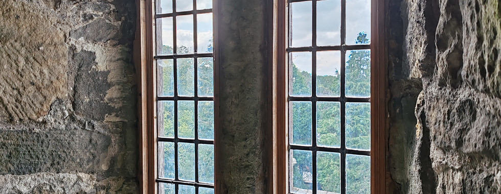 Rustic stone room with two tall, arched windows revealing a view of trees and rooftops. The setting is medieval, with an aged, historic feel.