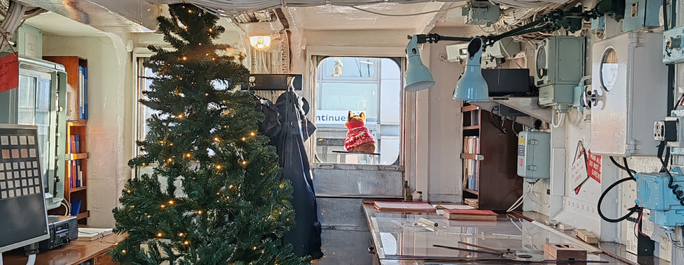 Inside a ship's cabin, a Christmas tree with lights stands amid wooden desks and equipment. A figure in a Santa hat is seen through a window.