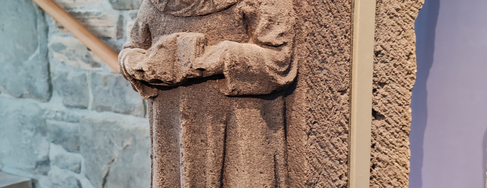 Stone sculpture of a person with curly hair in flowing attire, mounted on a textured back panel. Stone wall in the background.