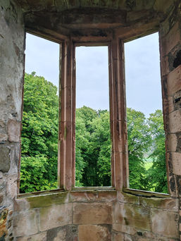 Stone window at Huntly Castle with three arches overlooks lush green trees. The weathered stone has a rustic charm against a cloudy sky.