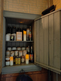 Pantry shelf with assorted jars and bottles, glowing candles, and a "Colman's Mustard" tin. Warm, vintage kitchen setting.