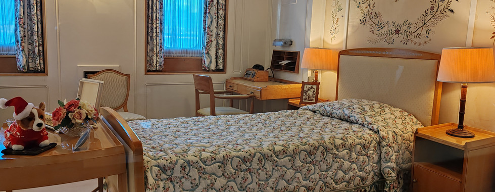 Elegant bedroom with floral bedding, wall art, and warm lighting. A stuffed dog toy dressed as Santa and flowers sit on a wooden table near a window.