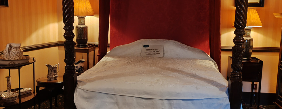 Elegant four-poster bed with rich red canopy in a vintage room with striped walls and floral carpet. A sign sits on the white bedding.