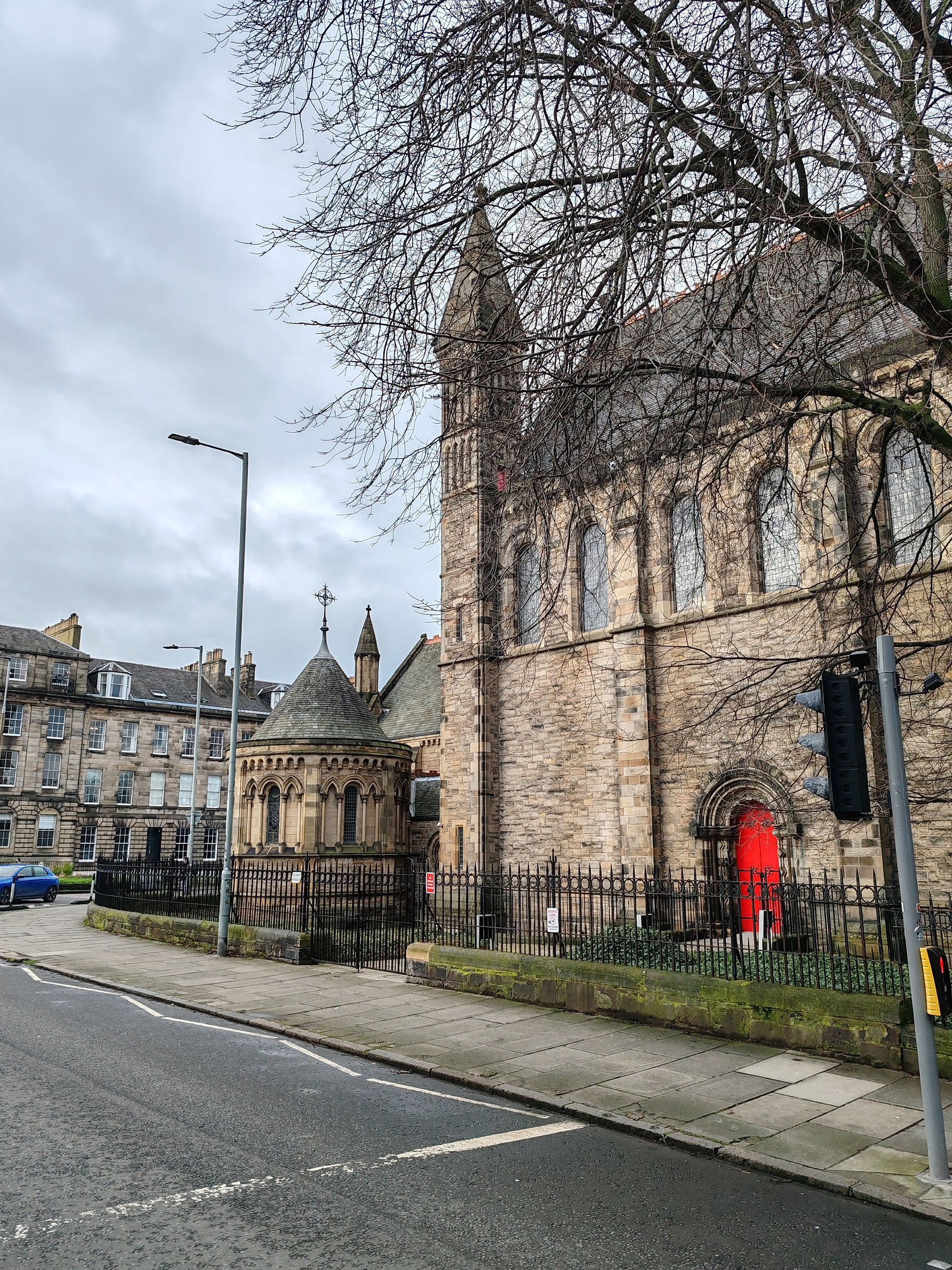 Historic stone church with a tall spire and red door, surrounded by bare trees. Gray sky and adjacent buildings. Quiet street scene.