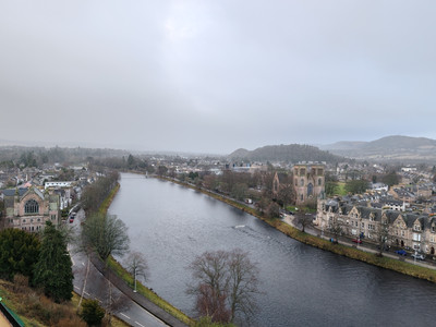 Aerial view of a town by the River Ness with historic stone buildings and Inverness Cathedral. Overcast sky, bare trees, and a peaceful winter scene.