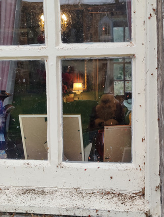 Window view into a cosy room with table lamps, framed pictures, a plant, curtains, and Santa Claus sitting in a chair. Warm lighting creates a homely atmosphere.