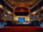 Ornate theater interior with red velvet seats, golden accents, and blue curtain backdrop. Empty stage, creating an elegant, grand atmosphere.