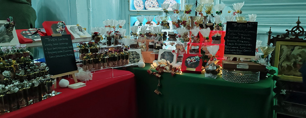 Tables with red and green cloths display truffle candies wrapped in colourful foil. Chalkboards list flavours. Elegant plates on shelves in background.