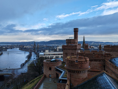 Scenic view of Inverness Castle's brick towers with a river winding through a city in the background, under a partly cloudy blue sky.