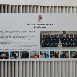 Sign titled "THE ROYAL YACHT BRITANNIA MAINTENANCE" with an image of a team in navy uniforms. Details about maintenance work and charitable trust info.