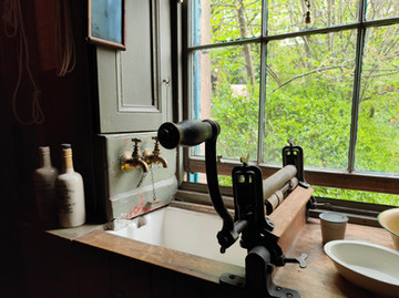 Old-fashioned mangle near a window with lush green trees outside, alongside a sink with brass faucets and vintage bottles.