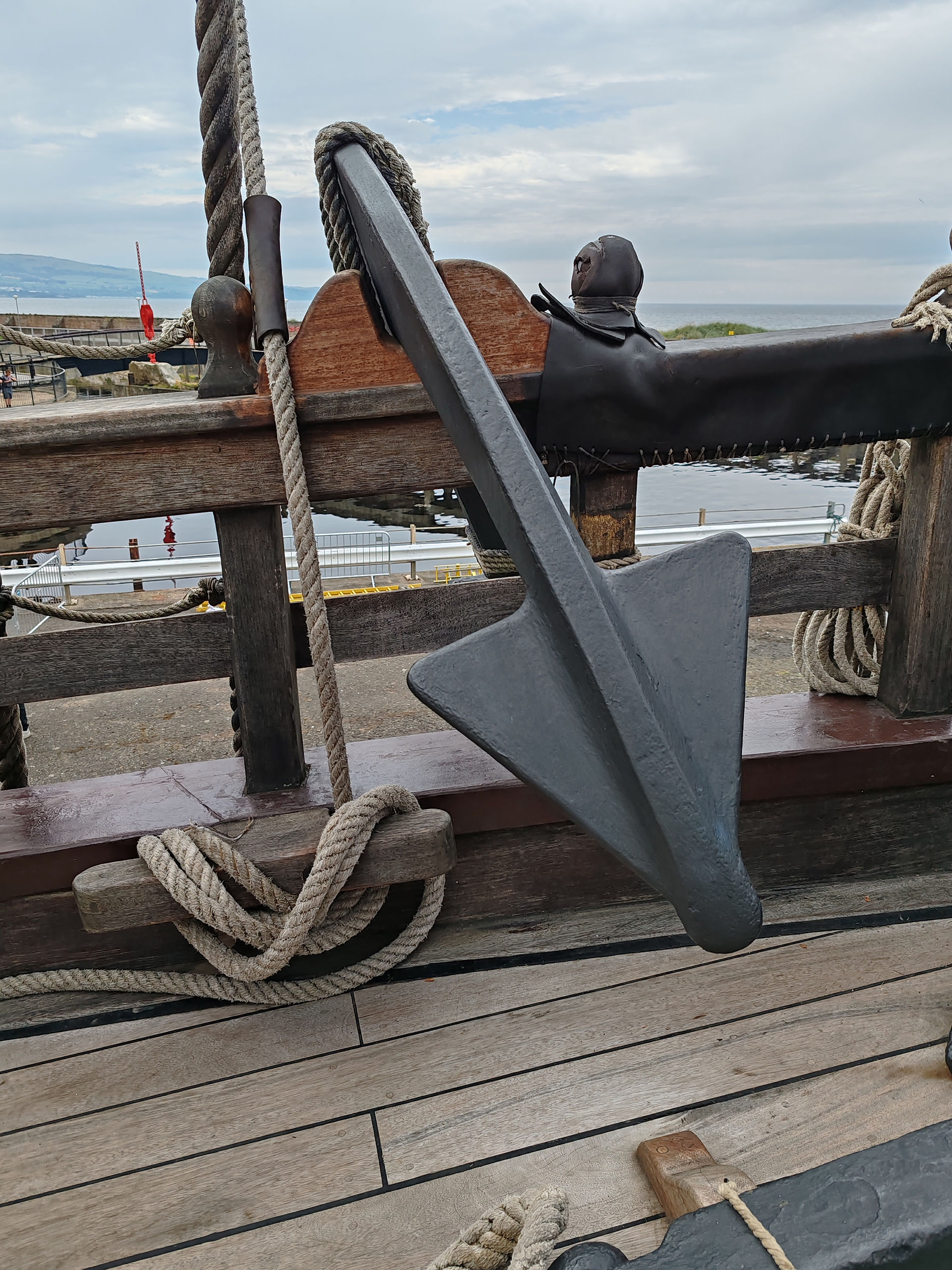 Large ship anchor and thick ropes on a wooden deck by the water, with a cloudy coastal harbour in the background.