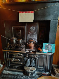 Vintage stove with kettles, pots, and a tea towel marked "L.N.E.R." in a cosy kitchen. An information card rests on the stovetop. Warm lighting.