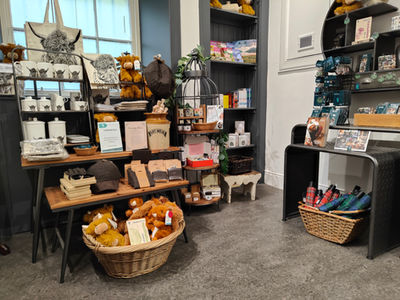 Shop display with highland-themed items. Shelves hold mugs, plush toys, books, and hats. A basket of umbrellas sits below. Cosy ambiance.