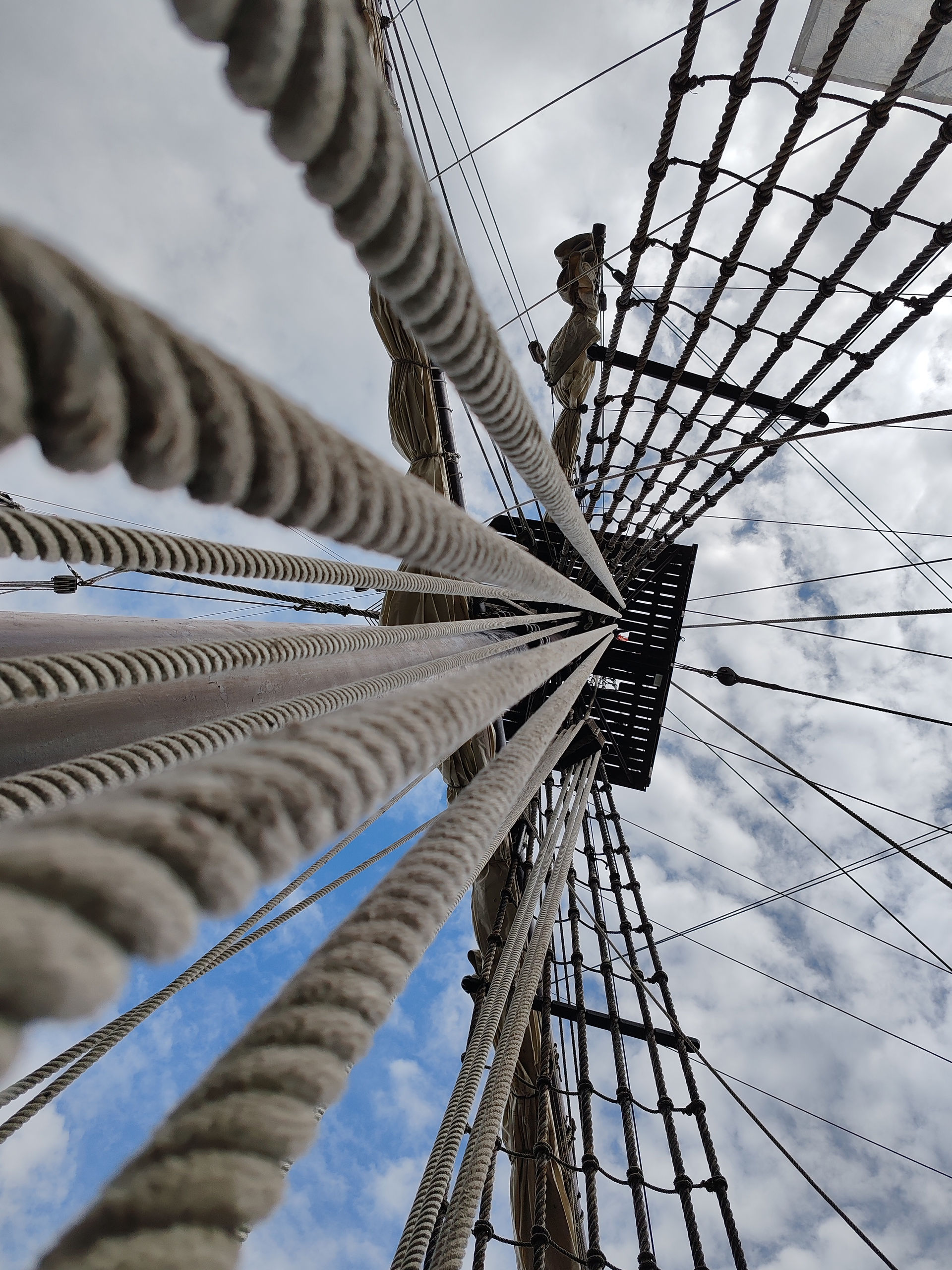 Upward view of a ship’s mast, rigging ropes, and netting against a cloudy sky, creating a dramatic nautical scene