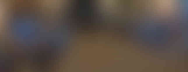 Floral-patterned sofas in a cosy, elegant room with a decorated fireplace and framed photos. Newspapers on a table, warm lighting.