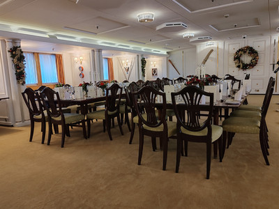 Elegant dining room with arranged tables and chairs, festive wreaths on walls, and soft lighting. White tablecloths, red floral centrepieces.