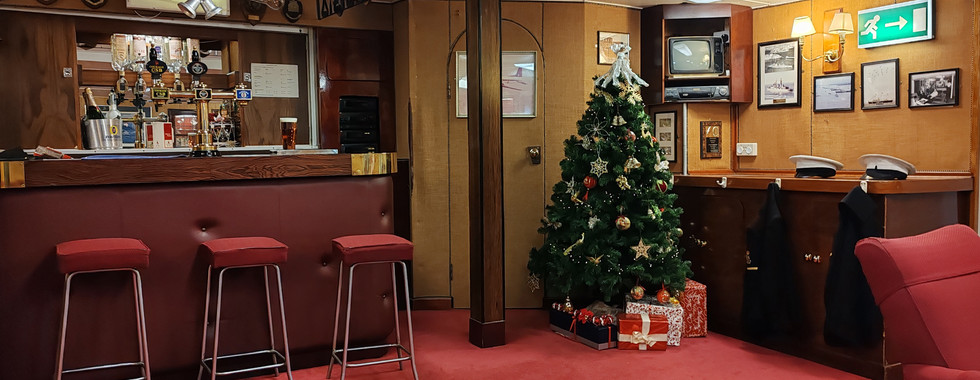 Cosy bar with a Christmas tree, red carpet, and three red stools. A decorated bar with drinks awaits. Gifts sit under the tree.