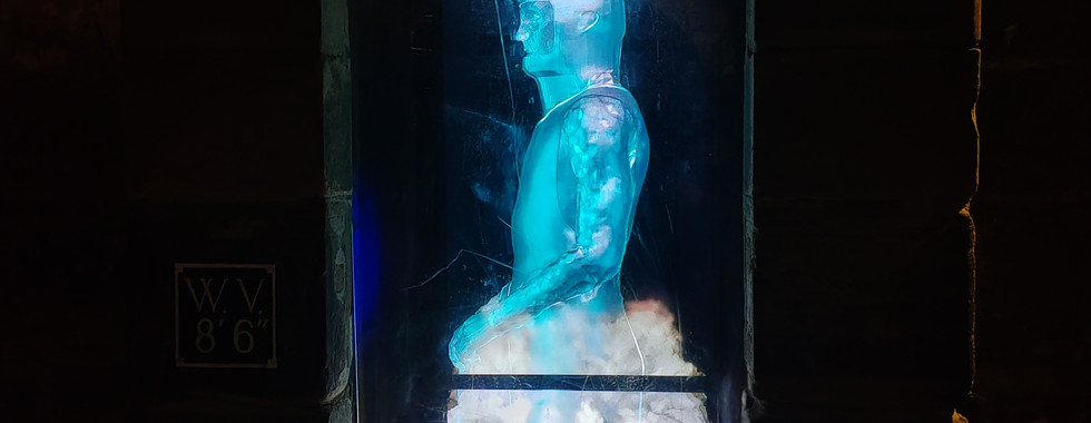 Glowing blue statue projection of a crowned figure in an arched niche. Surrounded by dark stone. Soft white clouds at the base. Mysterious mood.