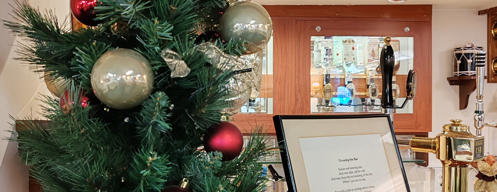 Small Christmas tree with red and silver ornaments on a wooden counter. Framed text next to a gold beer tap in a cosy bar setting.