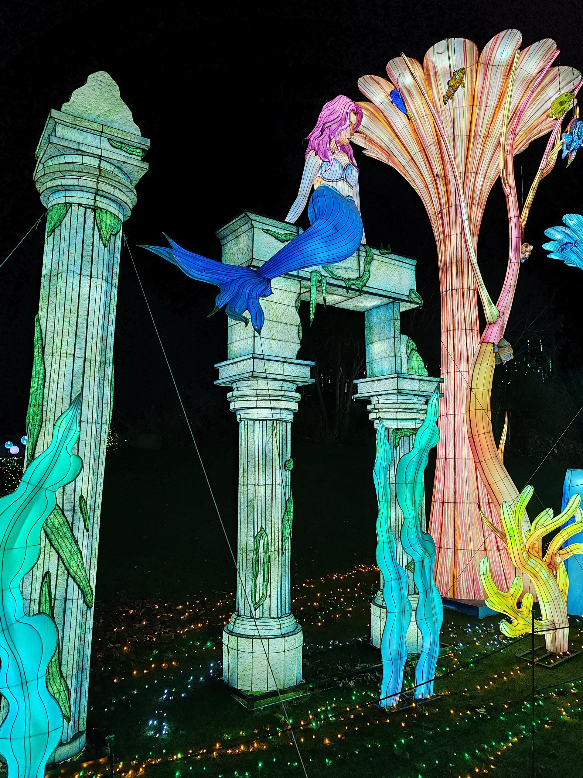A glowing mermaid on illuminated columns with seaweed, set against a dark backdrop. Bright coral structure and colourful underwater scene.