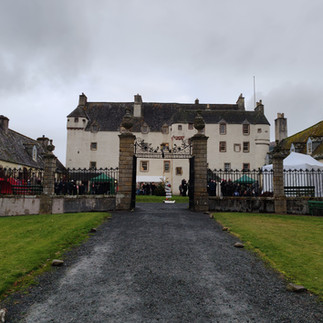 Historic white building, Traquair House, with a grey roof behind iron gates. People gather under tents, umbrellas visible. Overcast sky, green grass path.