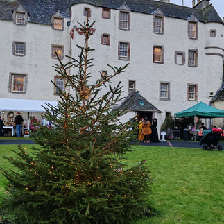 A lit Christmas tree stands on green grass before a historic building- Traquair House. People gather near stalls under canopies. It's overcast and festive.